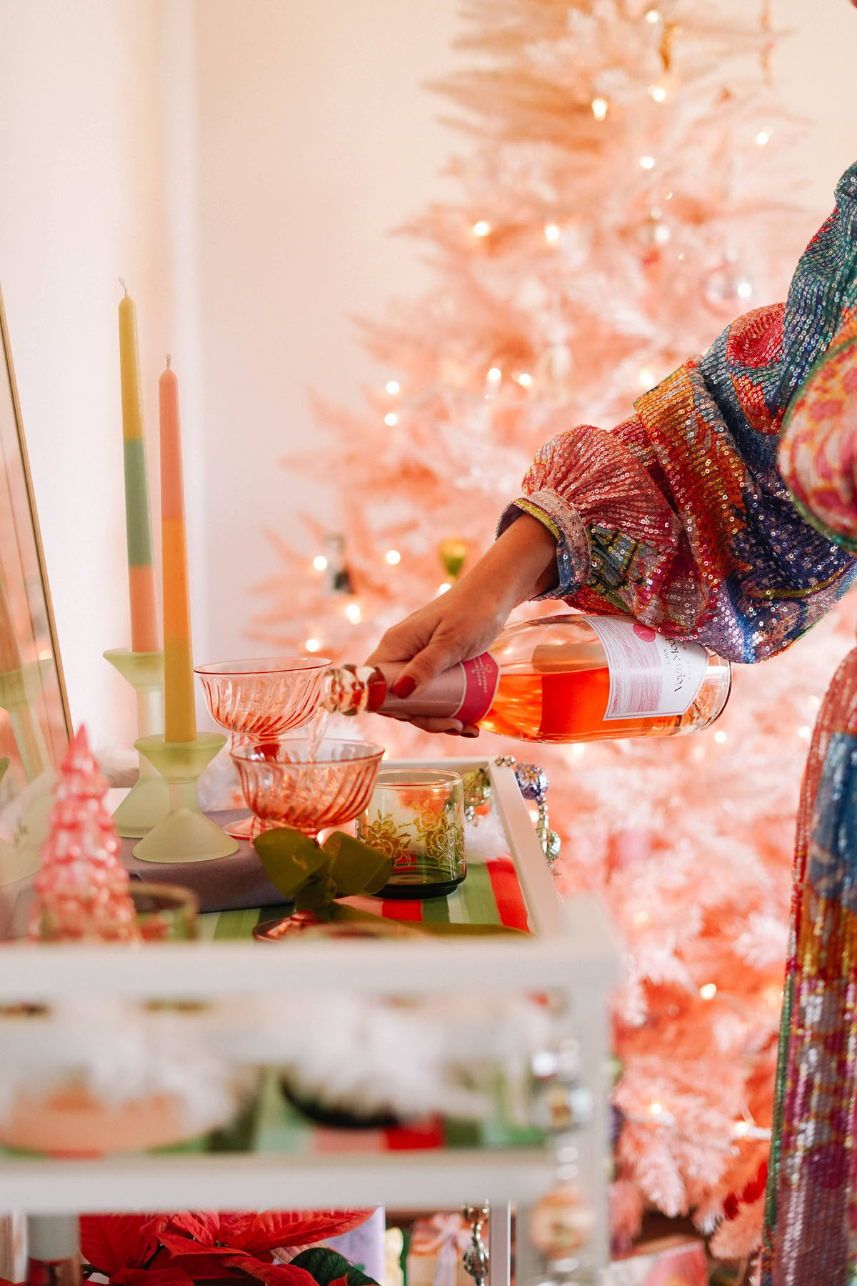 Pouring wine into pink champagne coupes in sparkly dress with pink christmas tree
